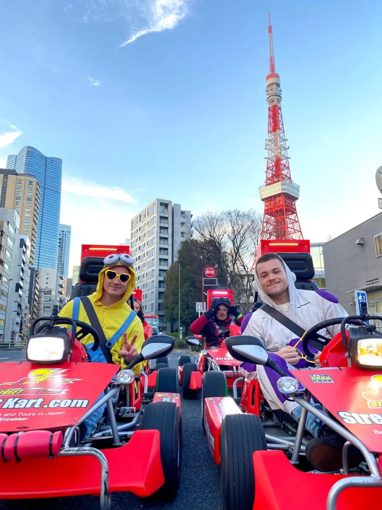 IMG_1084 - StreetKart People riding bright red go-karts in a city street, wearing colorful costumes (one in a yellow minion outfit with heart-shaped sunglasses) with Tokyo Tower visible in the background behind them.