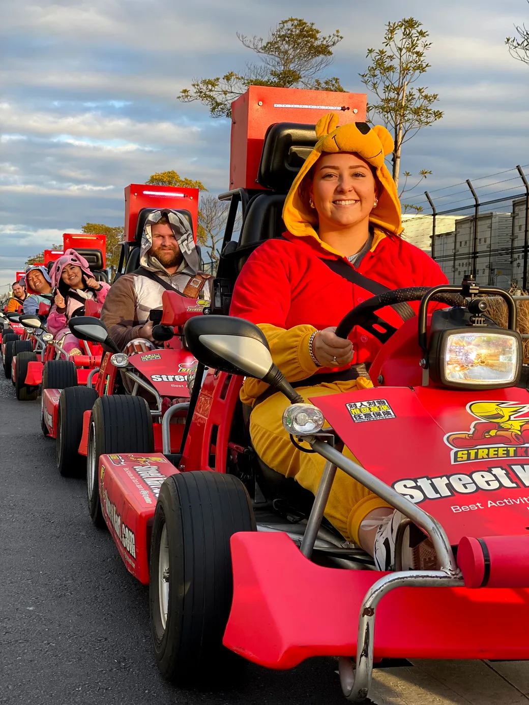 A group of people in bright costumes riding red go-karts in a line along an outdoor track, front rider wearing a bear hood smile on their face.