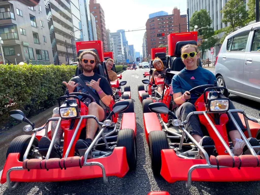 IMG_5257 - StreetKart A group of four people in red go-karts riding down a city street, smiling and wearing sunglasses.