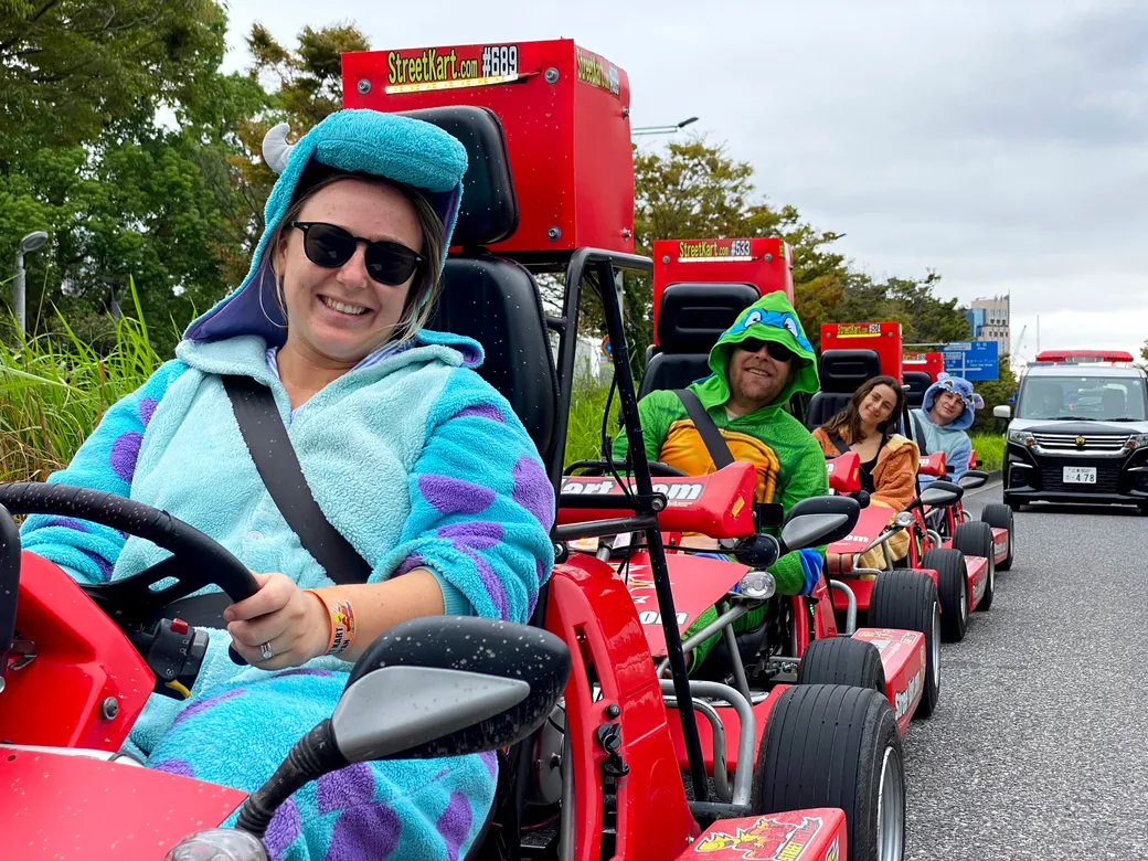 Person in a blue onesie driving a red go-kart, wearing sunglasses and a towel on head, smiling at camera on a parade-style line of go-karts.